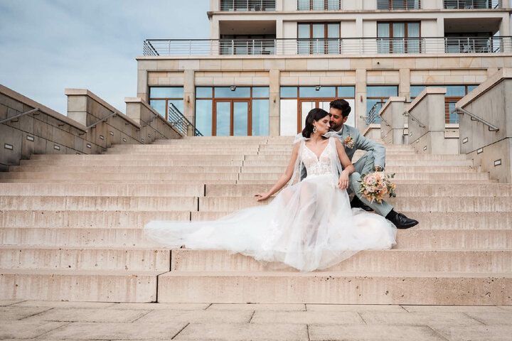 Brautpaar sitzt auf Treppe vor dem ATLANTIC Hotel Wilhelmshaven, modernes Gebäude im Hintergrund.