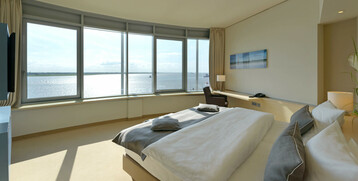 Bright hotel room with large bed and panoramic window overlooking the water.