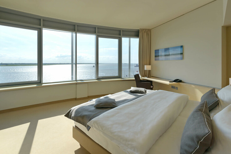 Bright hotel room with large bed and panoramic window overlooking the water.
