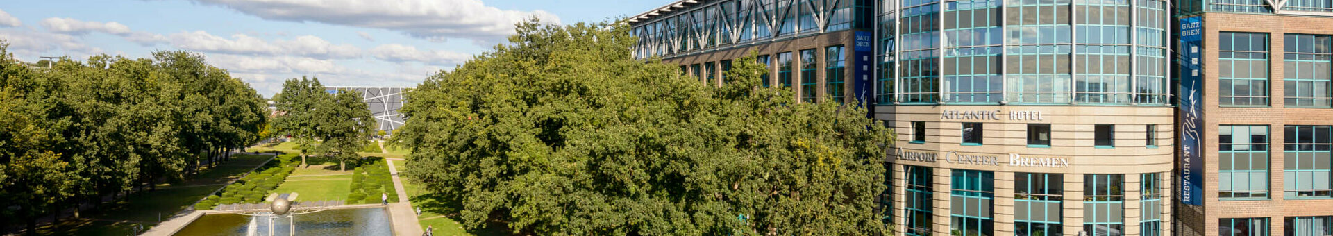 ATLANTIC Hotel Airport Bremen with modern glass façade, park with pond and trees in the foreground, blue sky.