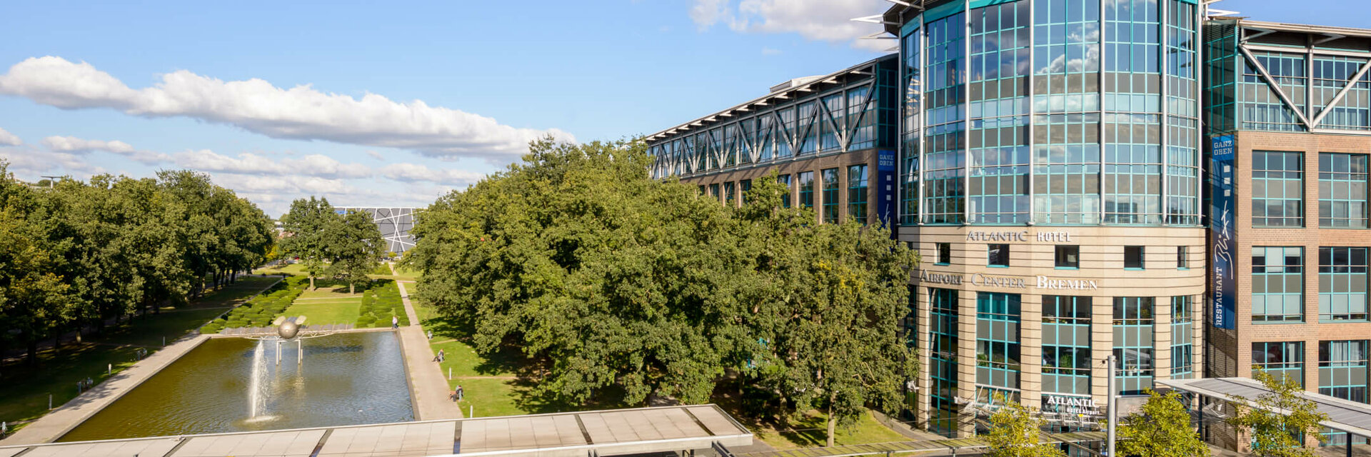 ATLANTIC Hotel Airport Bremen mit moderner Glasfassade, Park mit Teich und Bäumen im Vordergrund, blauer Himmel.