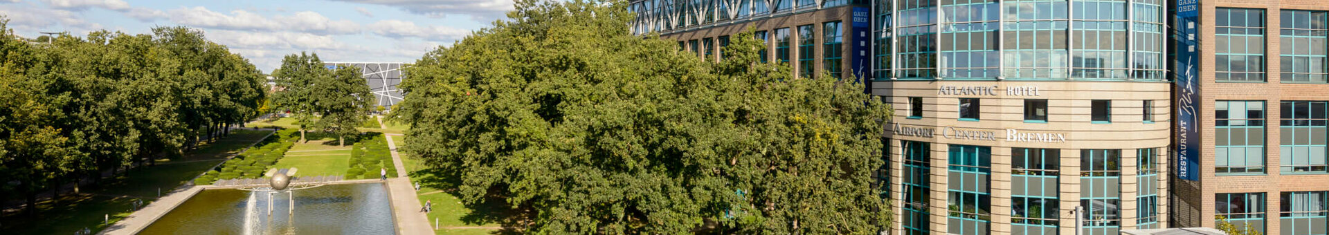 ATLANTIC Hotel Airport Bremen with modern glass façade, park with pond and trees in the foreground, blue sky.