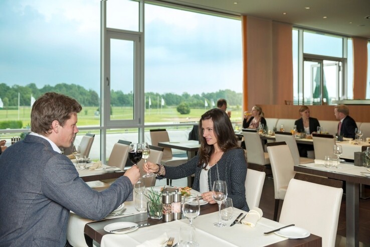 Hotelrestaurant mit Gästen, die an elegant gedeckten Tischen speisen. Große Fenster bieten Blick auf grüne Landschaft.