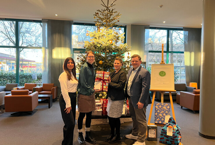 4 Personen stehen vor einem Weihnachtsbaum mit vielen Geschenken