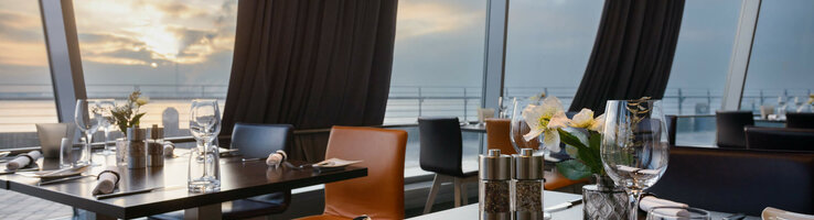 Gedeckte Tische im Restaurant STROM des ATLANTIC Hotel SAIL City Bremerhaven Modernes Restaurant mit gedeckten Tischen, großen Fenstern und Blick auf Sonnenuntergang über dem Wasser im ATLANTIC Hotel.