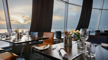 Set tables in the restaurant STROM in the ATLANTIC Hotel Sail City in Bremerhaven Modern restaurant with set tables, large windows and a view of the sunset over the water in the ATLANTIC Hotel.