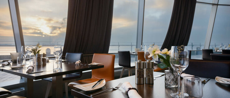 Set tables in the restaurant STROM in the ATLANTIC Hotel Sail City in Bremerhaven Modern restaurant with set tables, large windows and a view of the sunset over the water in the ATLANTIC Hotel.