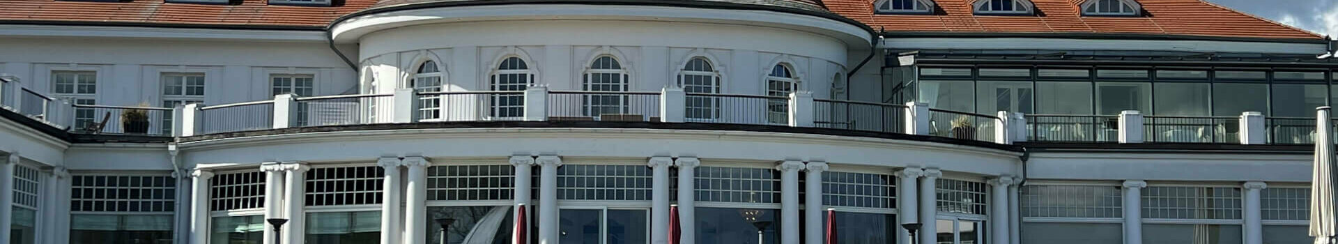 Historic hotel building with a red roof, two white cars in front of it on a well-tended lawn under a blue sky.