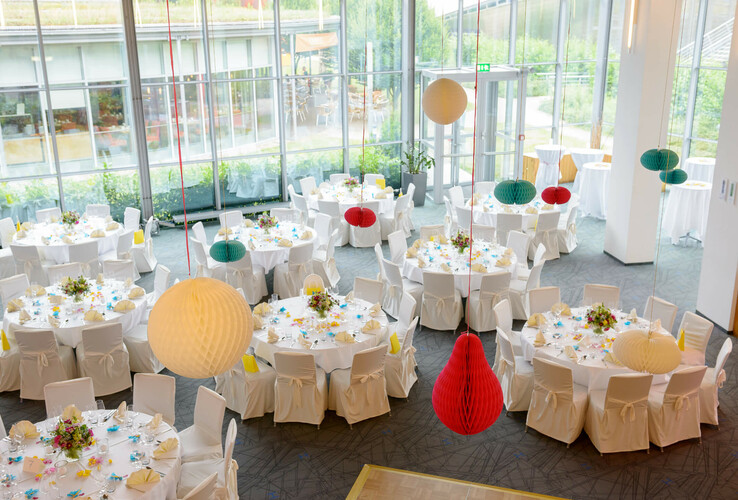 Großer, heller Bankettsaal mit runden, festlich gedeckten Tischen und farbigen Papierlampions.