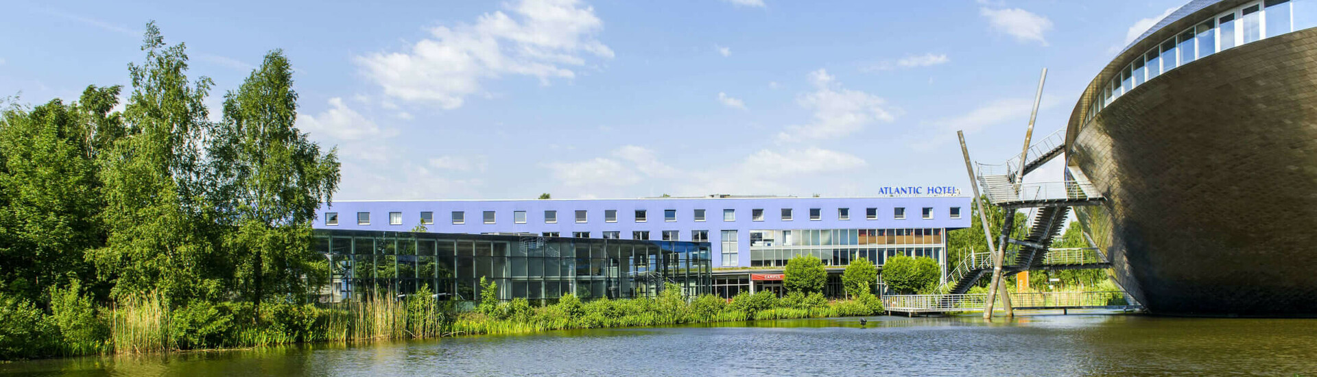 ATLANTIC Hotel Universum in Bremen Modernes Hotel am See, umgeben von Bäumen, mit blauem Himmel im Hintergrund.