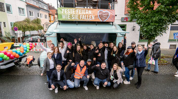 Gruppe fröhlicher Menschen in Freizeitkleidung posiert draußen vor einem bunt geschmückten Anhänger.