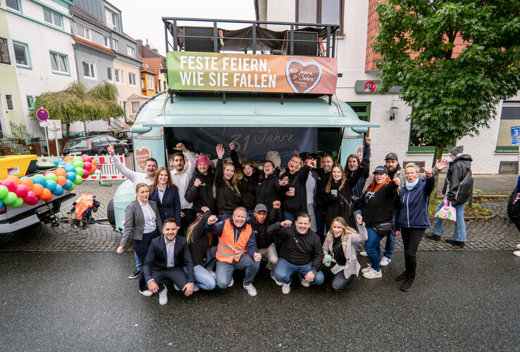 Gruppe fröhlicher Menschen in Freizeitkleidung posiert draußen vor einem bunt geschmückten Anhänger.
