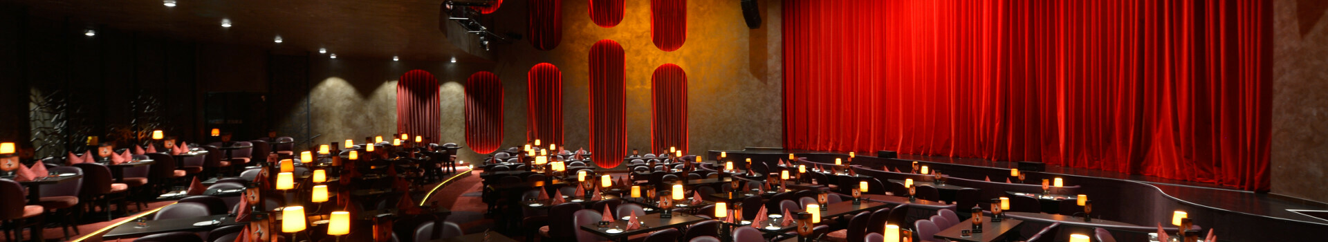 Elegantes Theater mit rotem Vorhang, gedämpfter Beleuchtung und Tischen, die mit Lampen und Servietten dekoriert sind.