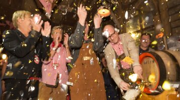 Menschen feiern in traditioneller Kleidung, während ein Mann ein Bierfass anschlägt, umgeben von Konfetti.