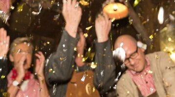 People celebrate in traditional dress while a man taps a beer keg, surrounded by confetti.