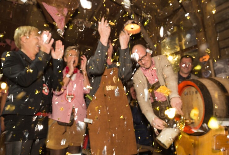 People celebrate in traditional dress while a man taps a beer keg, surrounded by confetti.