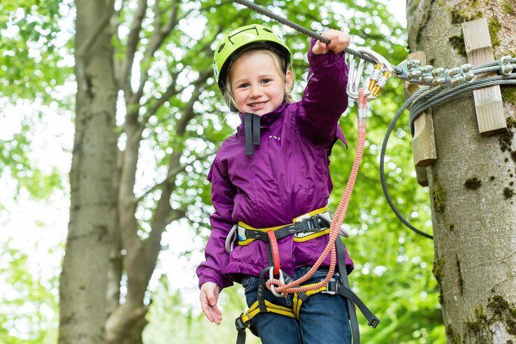 Kind klettert im Wald mit Sicherheitsausrüstung, lächelt fröhlich. Perfekt für Familienaktivitäten in Travemünde.