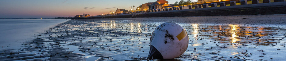 Strand bei Sonnenuntergang mit Boje im Vordergrund und beleuchtetem Hotel im Hintergrund.
