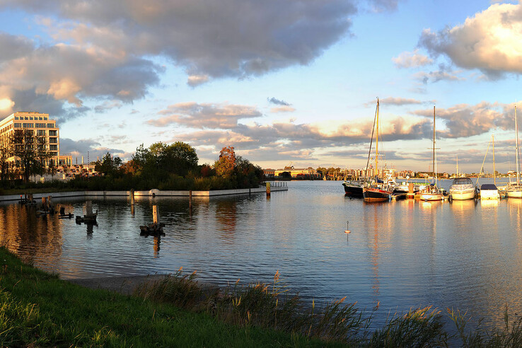 Hotel am Wasser mit Segelbooten im Hafen, umgeben von Bäumen, unter bewölktem Himmel bei Sonnenuntergang.
