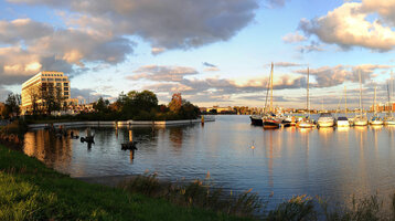 Hotel am Wasser mit Segelbooten im Hafen, umgeben von Bäumen, unter bewölktem Himmel bei Sonnenuntergang.