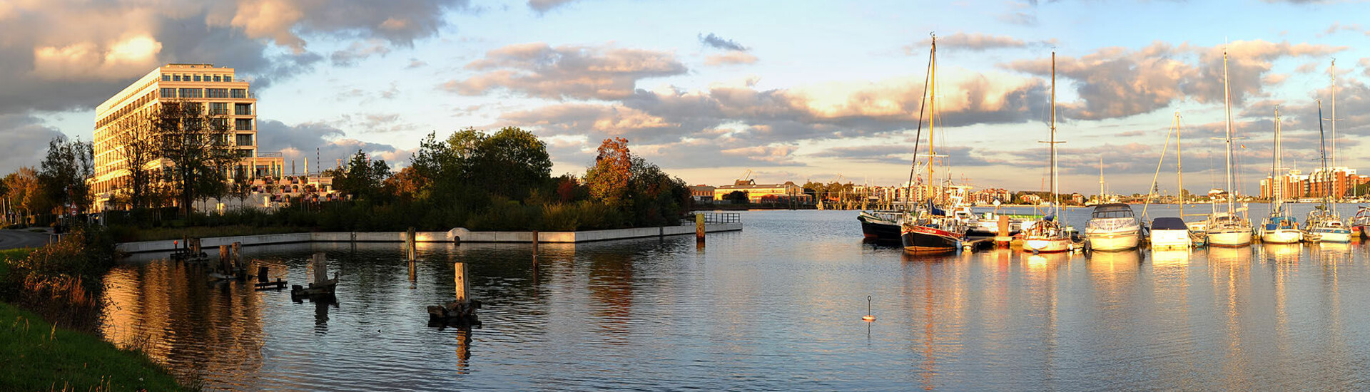 Außenansicht ATLANTIC Hotel Wilhelmshaven Hotel am Wasser mit Segelbooten im Hafen, umgeben von Bäumen, unter bewölktem Himmel bei Sonnenuntergang.