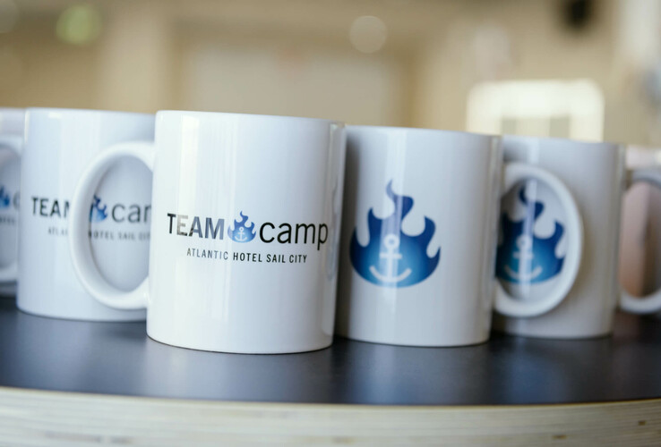 White cups with logo and lettering "TEAMcamp, ATLANTIC Hotel Sail City" arranged on a table.