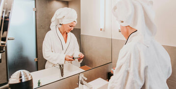 Frau im Bademantel mit Handtuchturban im modernen Hotelbad des ATLANTIC Hotel Kiel, vor einem Spiegel stehend.
