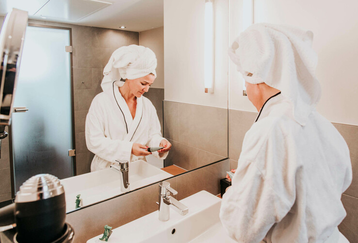 Woman in bathrobe with towel turban in the modern hotel bathroom of the ATLANTIC Hotel Kiel, standing in front of a mirror.