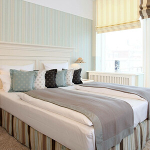 Helles Hotelzimmer mit Doppelbett, gestreiften Kissen, Nachttischlampen und großen Fenstern mit gestreiften Vorhängen.