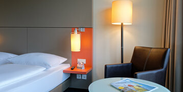 Modern hotel room with bed, bedside table, floor lamp, leather armchair and glass table with magazine.
