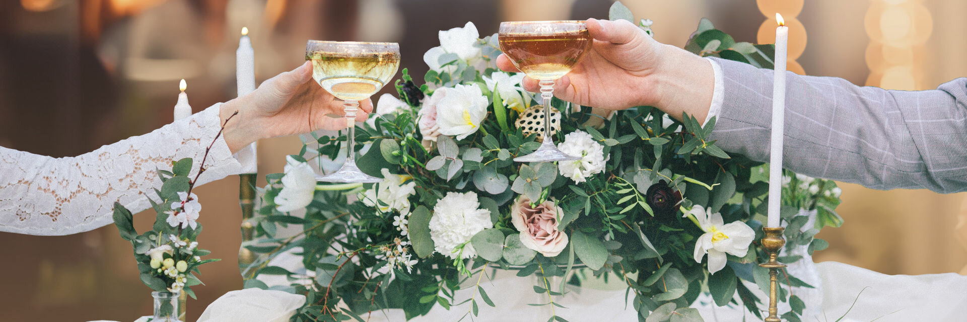 Zwei Personen stoßen mit Champagnergläsern an, vor Blumenarrangement und Kerzen.