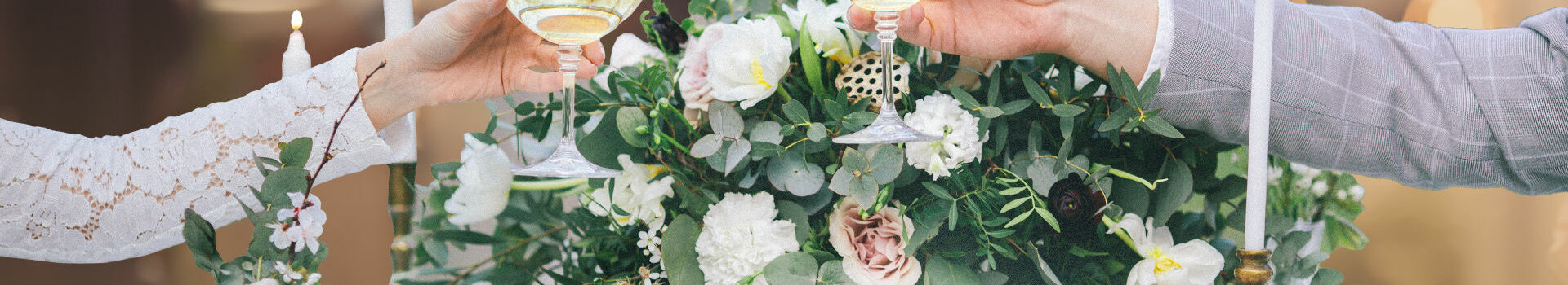 Zwei Personen stoßen mit Champagnergläsern an, vor Blumenarrangement und Kerzen.
