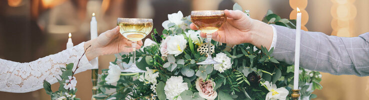Zwei Personen stoßen mit Champagnergläsern an, vor Blumenarrangement und Kerzen.