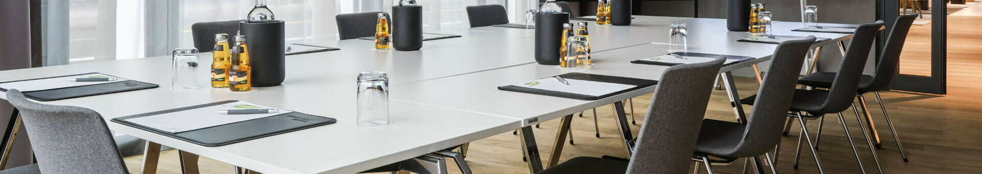 Moderner Konferenzraum mit großem Tisch, Stühlen, Schreibmaterial, Wasserflaschen und natürlichem Licht im ATLANTIC Hotel Münster.