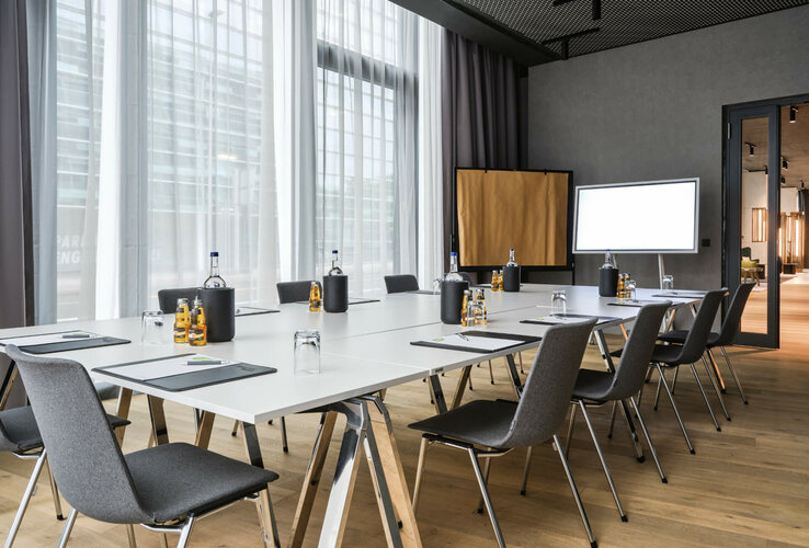 Moderner Konferenzraum mit großem Tisch, Stühlen, Schreibmaterial, Wasserflaschen und natürlichem Licht im ATLANTIC Hotel Münster.