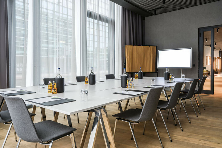 Moderner Konferenzraum mit großem Tisch, Stühlen, Schreibmaterial, Wasserflaschen und natürlichem Licht im ATLANTIC Hotel Münster.