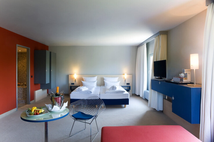 Modernes Hotelzimmer mit Doppelbett, stilvollen Möbeln, TV, Sessel und kleinem Tisch mit Sekt und Obst.