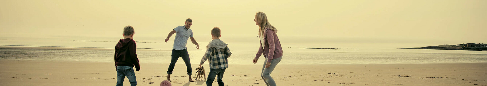 Familie spielt mit Hund und Ball am sonnigen Strand, Meer im Hintergrund.