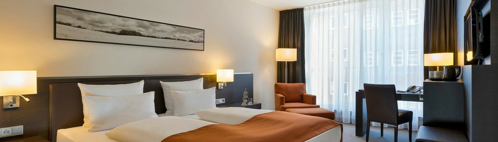 Modernes Hotelzimmer mit Doppelbett, orangefarbener Decke, Schreibtisch, Sessel und großen Fenstern mit Vorhängen.