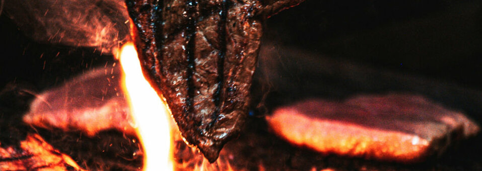 Ein saftiges Steak wird mit einer Zange über einer offenen Flamme auf dem Grill gehalten.