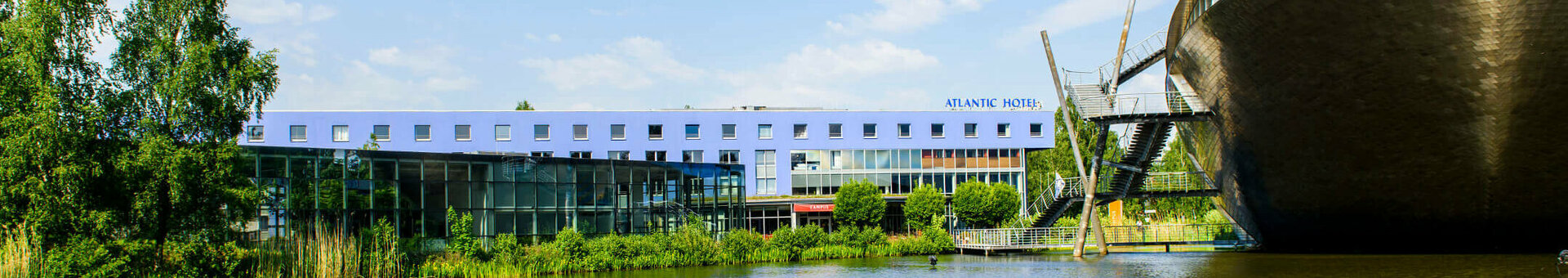 Aussenaufnahme Seeseite | ATLANTIC Hotel Universum in Bremen ATLANTIC Hotel Universum in Bremen neben einem See, moderner Bau, blauer Himmel, grüne Ufervegetation.