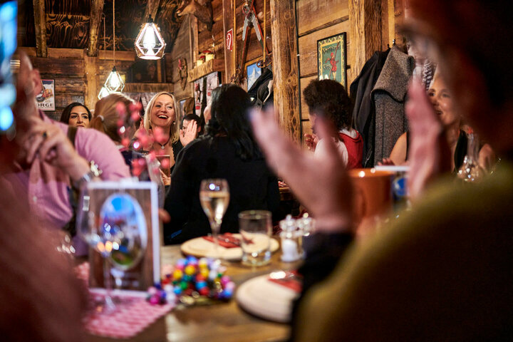 Apres ski party atmosphere in the ATLANTIC Almhütte Cozy restaurant atmosphere in the ATLANTIC Grand Hotel Bremen, with smiling guests and decorative wooden furniture.