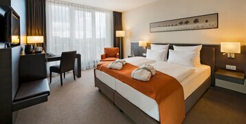 Modernes Hotelzimmer mit Doppelbett, orangefarbenem Überwurf, Schreibtisch, Sessel und großen Fenstern im ATLANTIC Hotel Lübeck.