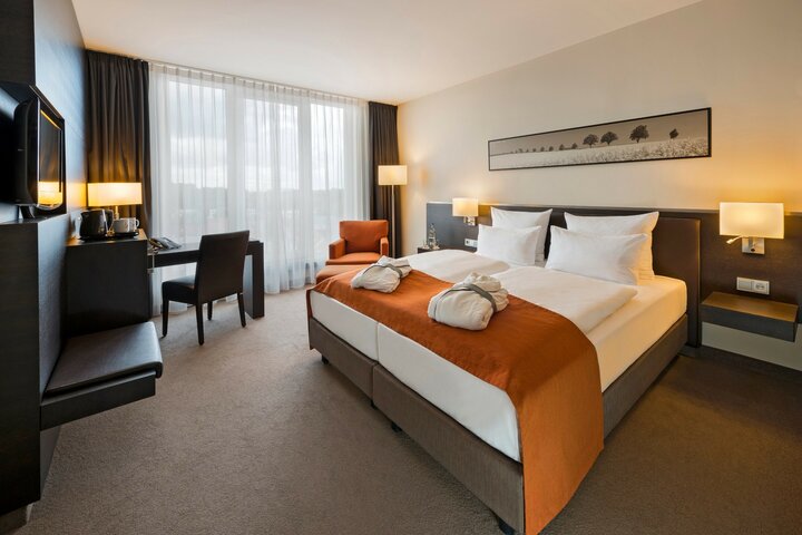 Modernes Hotelzimmer mit Doppelbett, orangefarbenem Überwurf, Schreibtisch, Sessel und großen Fenstern im ATLANTIC Hotel Lübeck.