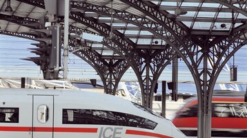 Moderner Bahnhof mit ICE-Zug im Vordergrund und einem weiteren Zug im Hintergrund unter einem gewölbten Glasdach.