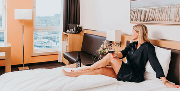 Frau entspannt auf Hotelbett im ATLANTIC Hotel Kiel, mit Weinglas, Blick auf Hafen durch Fenster.