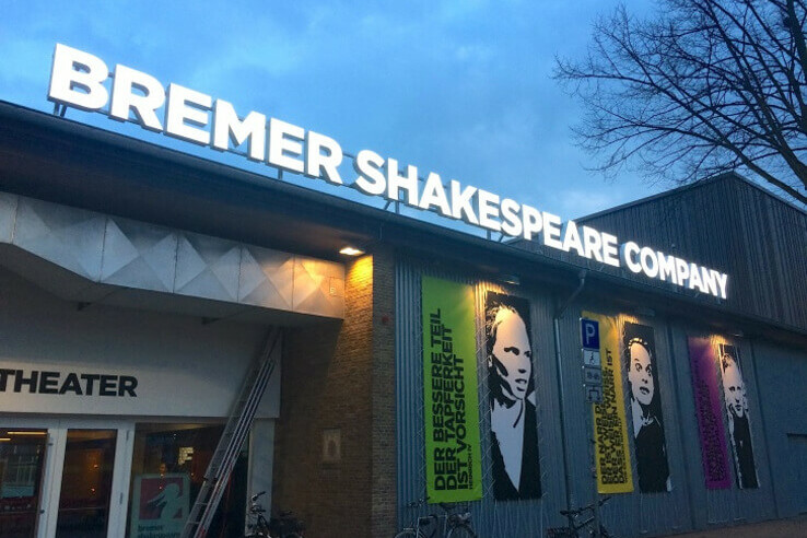 Theater façade with "Bremer Shakespeare Company" lettering and colorful posters, evening atmosphere.