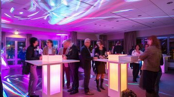 Eine Gruppe von Menschen steht an beleuchteten Stehtischen in einem farbig erleuchteten Veranstaltungsraum.