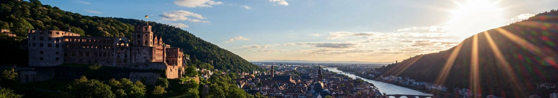 Blick auf Schloss und Stadt am Fluss bei Sonnenuntergang, umgeben von grünen Hügeln.