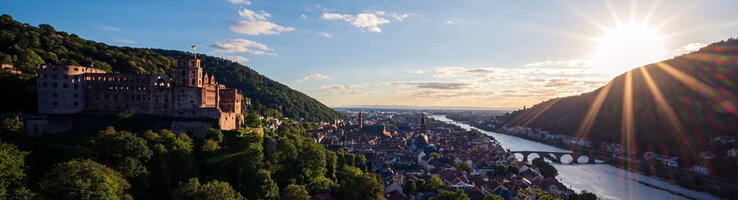 Blick auf Schloss und Stadt am Fluss bei Sonnenuntergang, umgeben von grünen Hügeln.